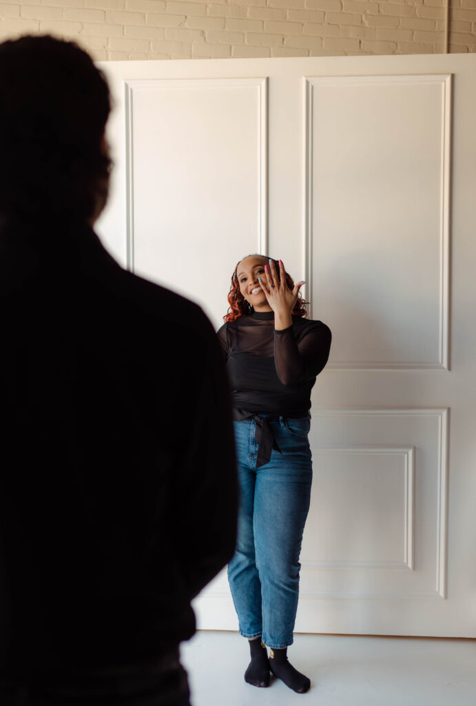 Engaged woman smiling while showing her engagement ring as her fiancé watches during their studio session in Augusta, Georgia