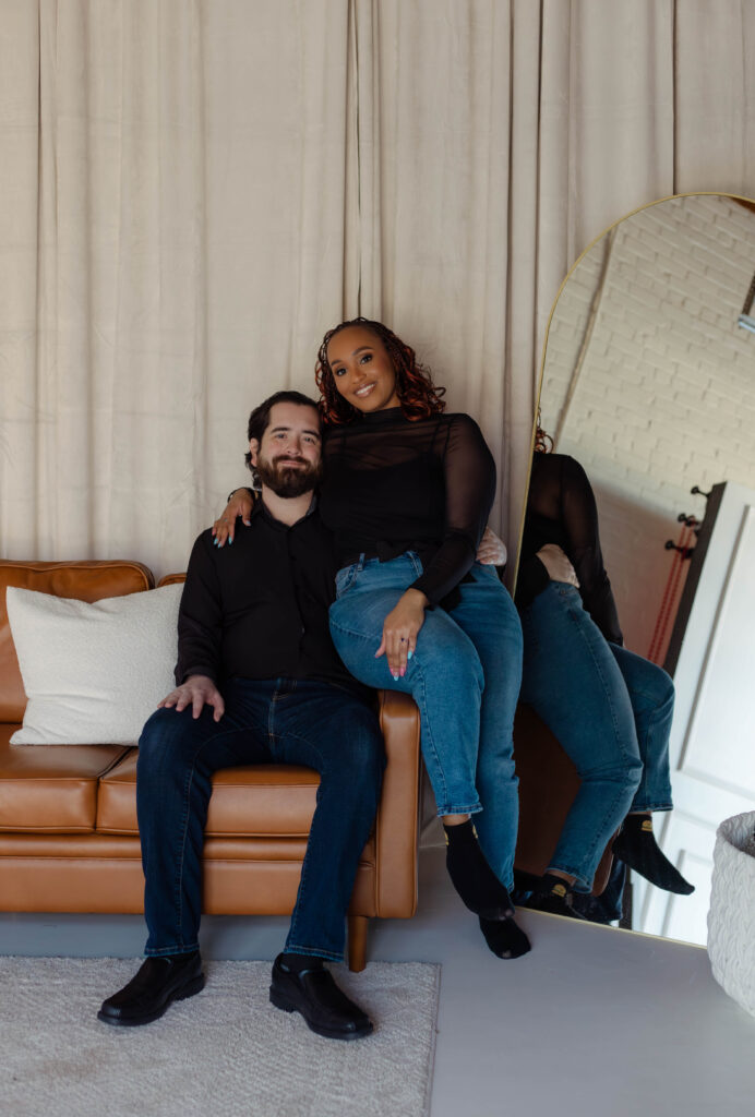 Engaged couple poses on a light brown leather couch during their studio engagement session in Augusta, Georgia
