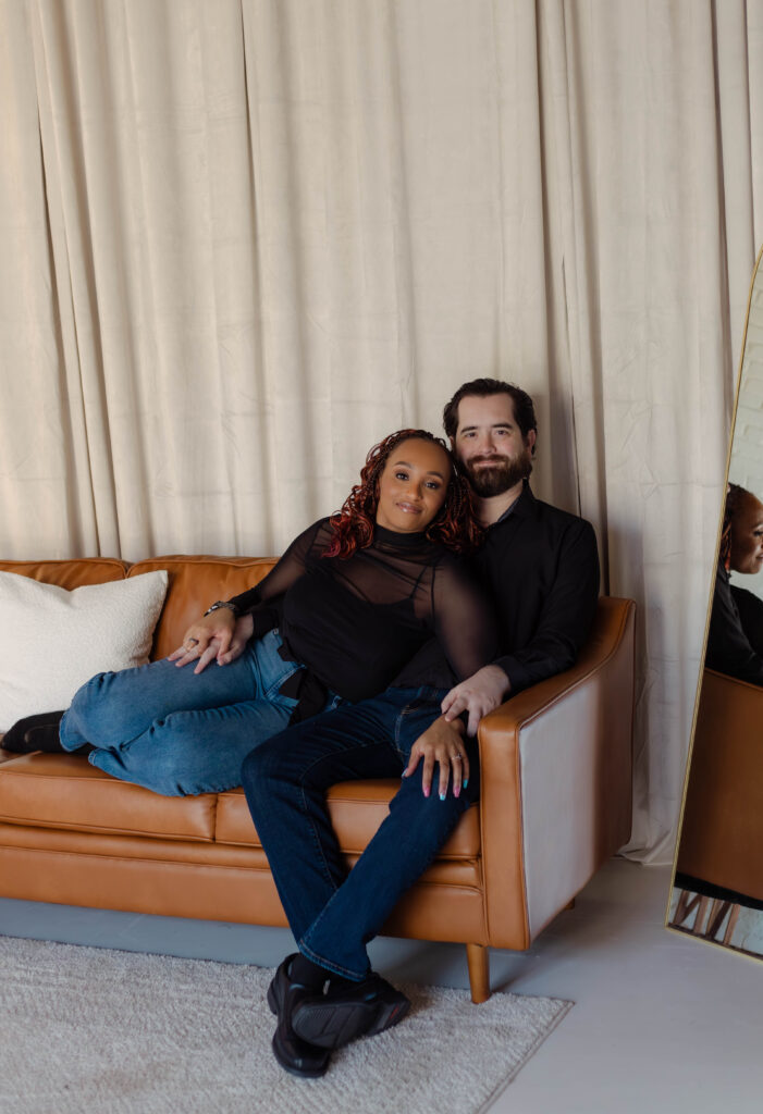 Engaged couple poses on a light brown leather couch during their photoshoot at Opal Bay Studio in Augusta, Georgia