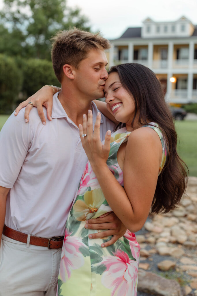 Man kisses his fiancée on the forehead while she shows off the engagement ring