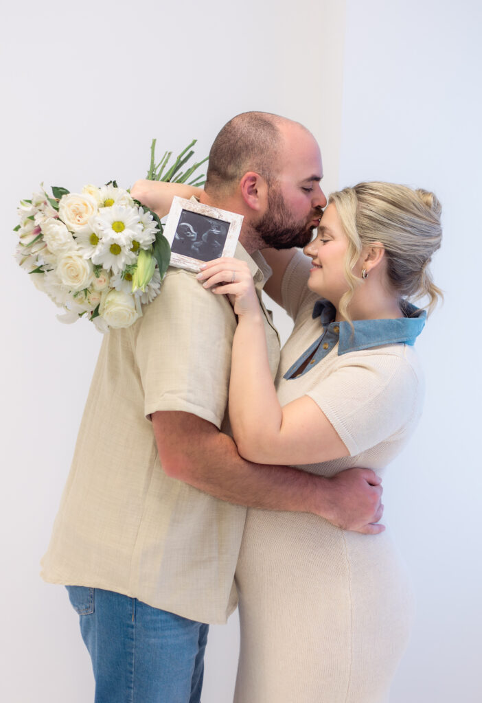 Husband kisses wife's forehead while she holds flowers and framed ultrasound photo