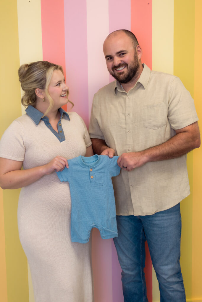 Couple holds blue onesie while standing in front of colorful striped wall