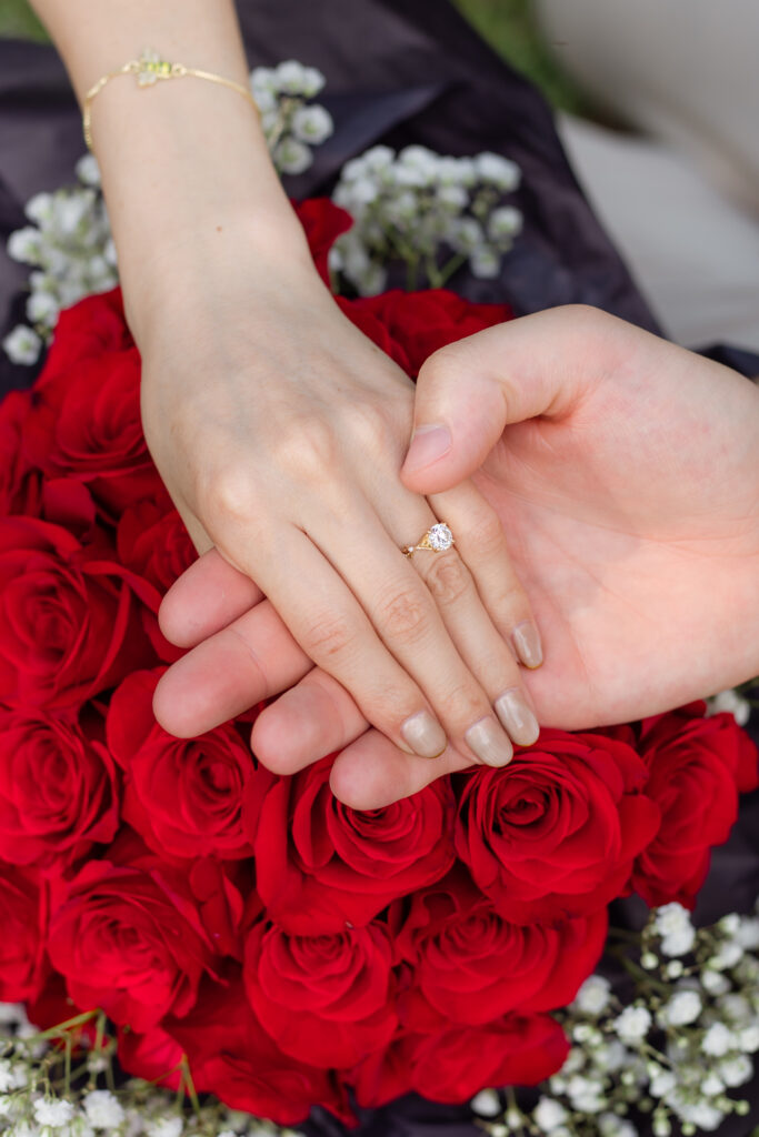 An up close photo of a woman's ring while husband holds her hand on some red roses