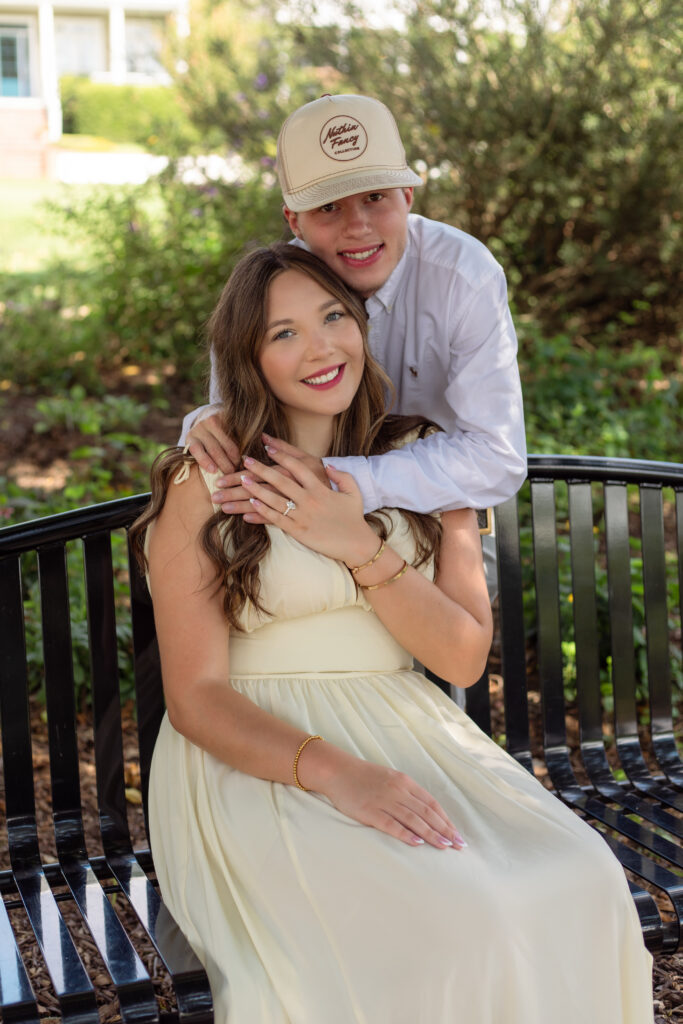Woman sits on bench while fiancé hugs her from behind